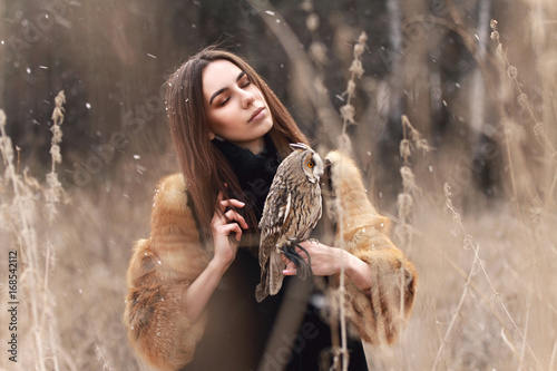 Woman in fur coat with owl on hand by first autumn snow. Beautiful brunette with long hair in nature, holding an owl. Romantic, delicate look girls