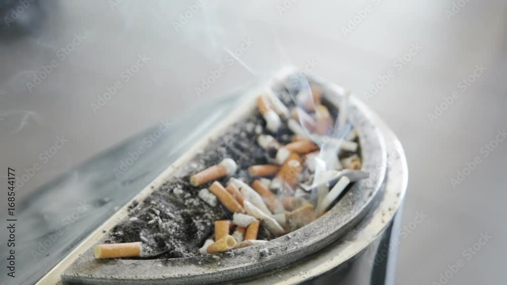 Smoldering cigarette butts in a large metal ashtray, full of ashes. Smoldering cigarette butts in a large metal ashtray, full of ashes.