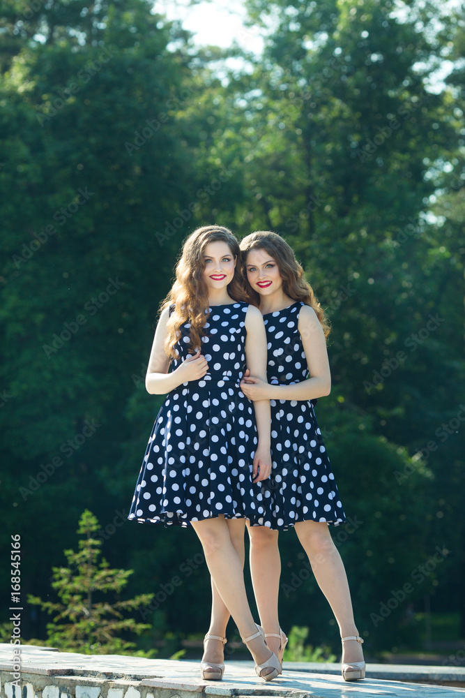 Two beautiful smiling girl sisters twins for a walk in summer park on a background of greenery with makeup and hairstyle