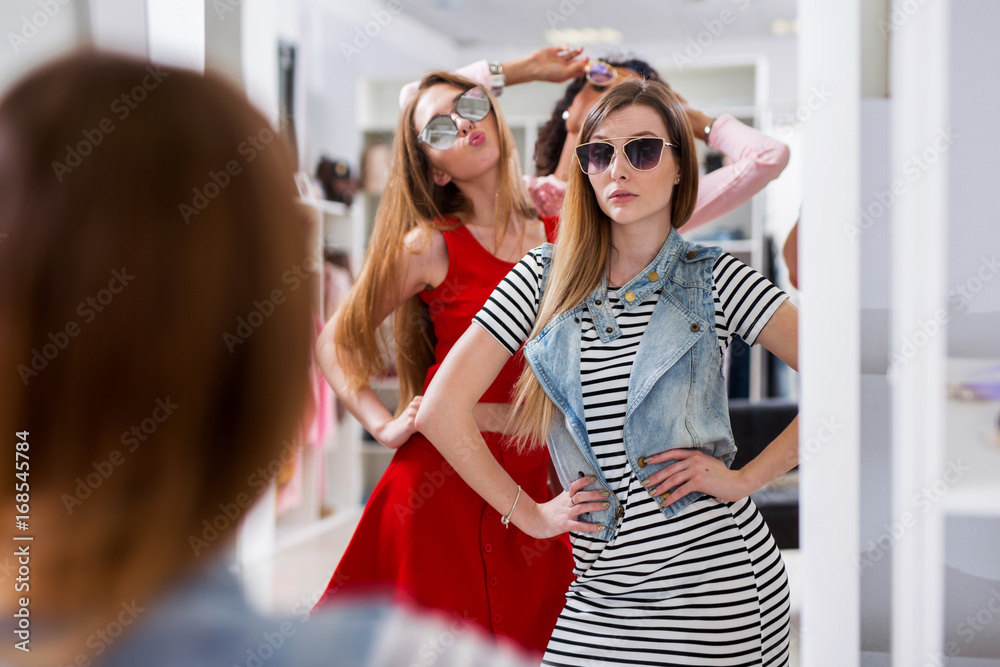 Glamorous girls trying on sunglasses posing in front of the mirror in fashion boutique Glamorous girls trying on sunglasses posing in front of the mirror in fashion boutique