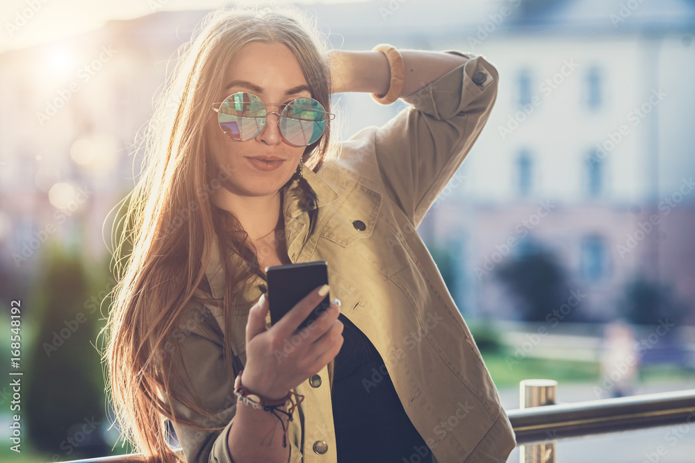 Fototapeta premium Young stylish pretty Woman, hands holding a phone. Sunset background, Sunny day,good weather, sunglasses, cool accessories.