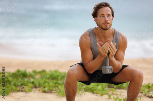 Fototapeta Naklejka Na Ścianę i Meble -  Fitness man training squat with dumbbell weight doing exercise for glutes muscles and quads, working out legs on beach.