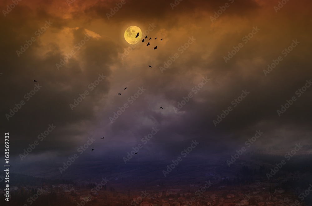 Full moon over the city . The yellow moon over the city . Look with top of a roof of houses . hilly terrain . The village in mountains . Silhouette of birds and lunar sky