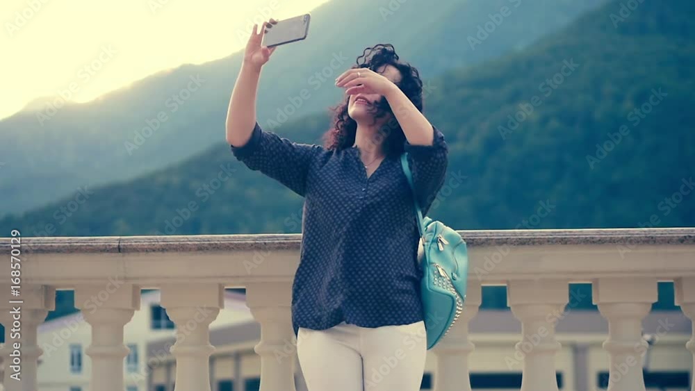 Smart, beautiful brunette corrects curly hair and talks on the phone by Skype on the background of outstanding mountains and beautiful sunset.