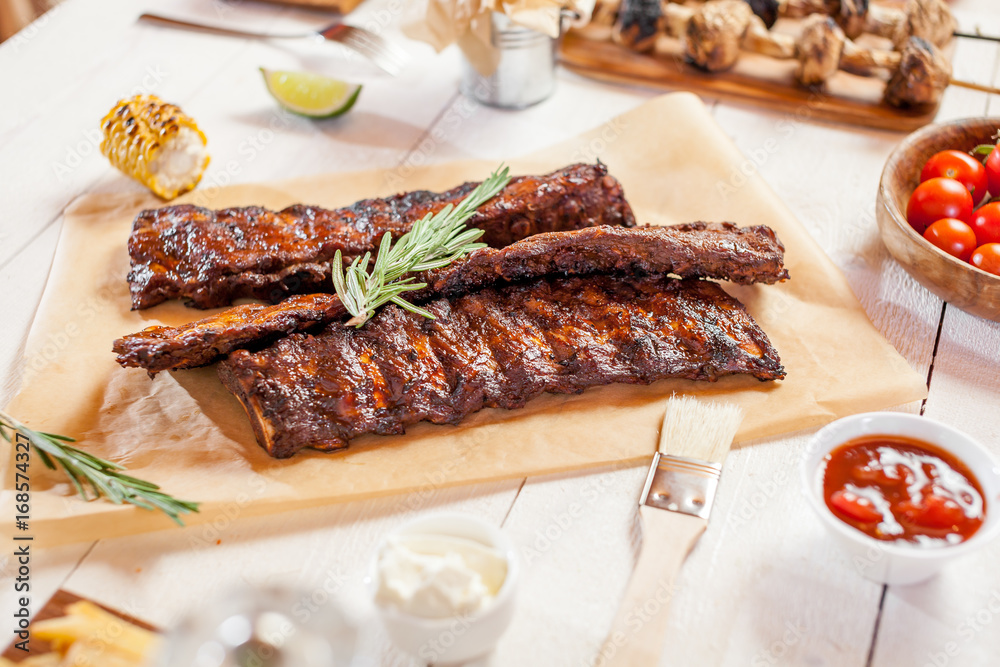 Spicy hot grilled spare ribs from a summer BBQ served with chips, corn and fresh tomatoes on an old wooden cutting board