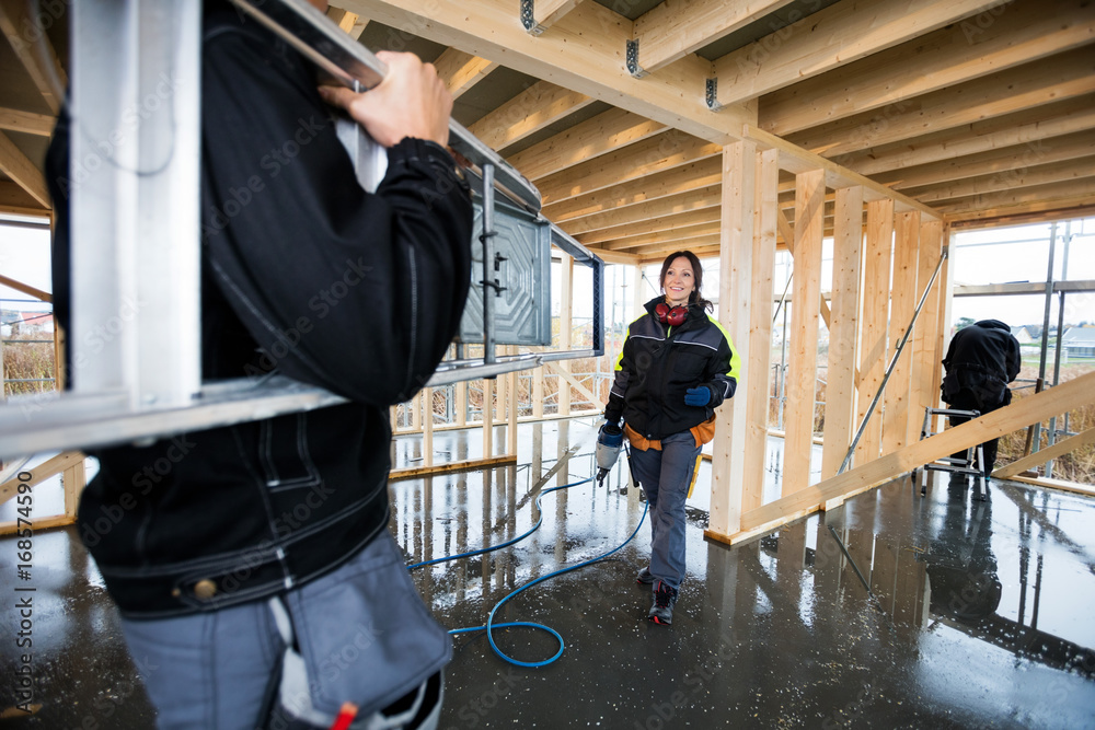 Female Carpenter Smiling While Male Colleague Carrying Ladder