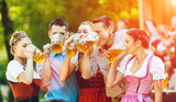 Im Biergarten - Freunde in Lederhosen und Dirndl stehen vor Blaskapelle mit Bierkrügen