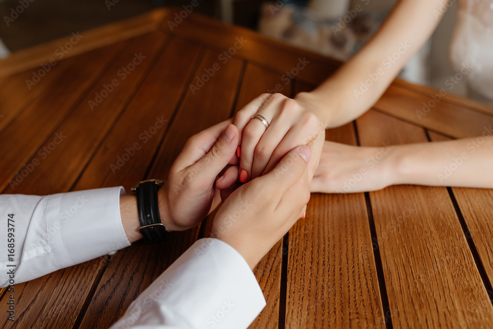 Bride and groom sitting at the table and holding hands on wooden background at wedding day. Close-up. Wedding ring. Bride and groom sitting at the table and holding hands on wooden background at wedding day. Close-up. Wedding ring.
