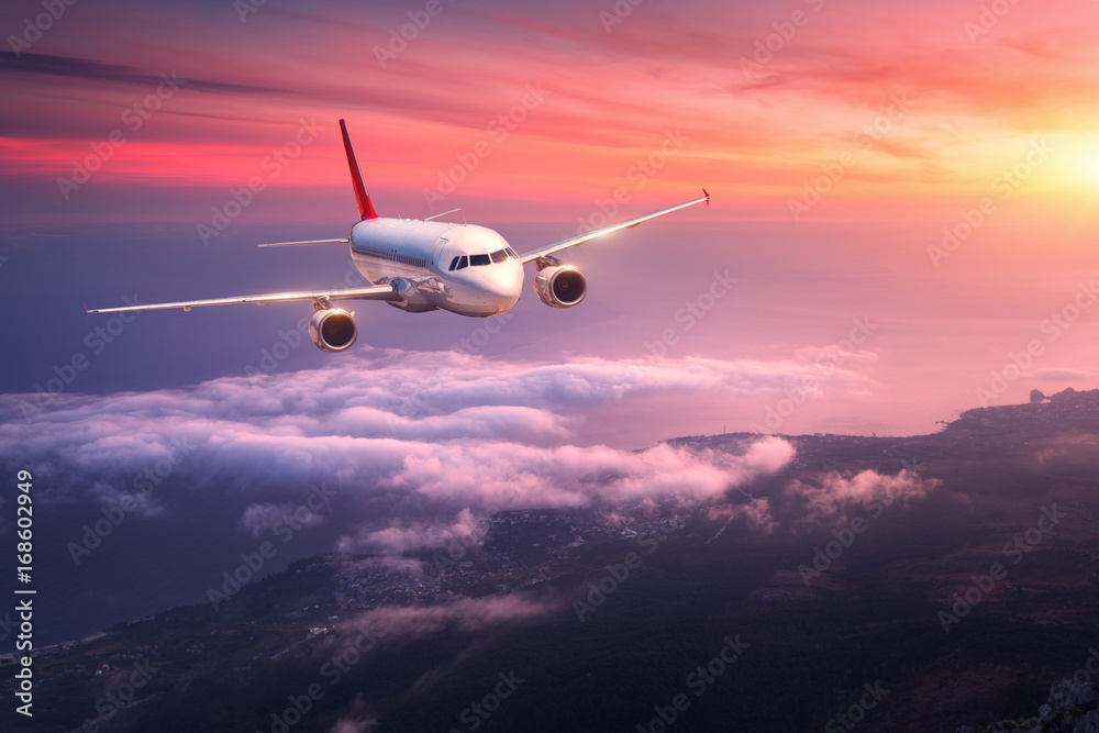 Naklejka premium Samolot pasażerski. Krajobraz z dużym białym samolotem lata w czerwonym niebie nad chmurami i morzem przy kolorowym zmierzchem. Samolot pasażerski ląduje o zmierzchu. Podróż służbowa. Samolot komercyjny. Podróż