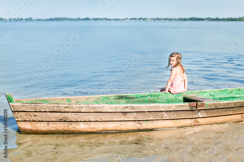 Portrait happy little girl in swimsuit sits in a boat near the shore of lake. Back view photo