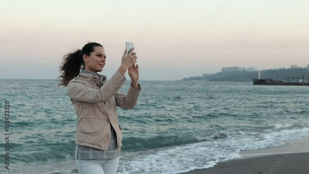 Young woman in a beige jacket takes pictures of herself on the beach in the evening Young woman in a beige jacket takes pictures of herself on the beach in the evening
