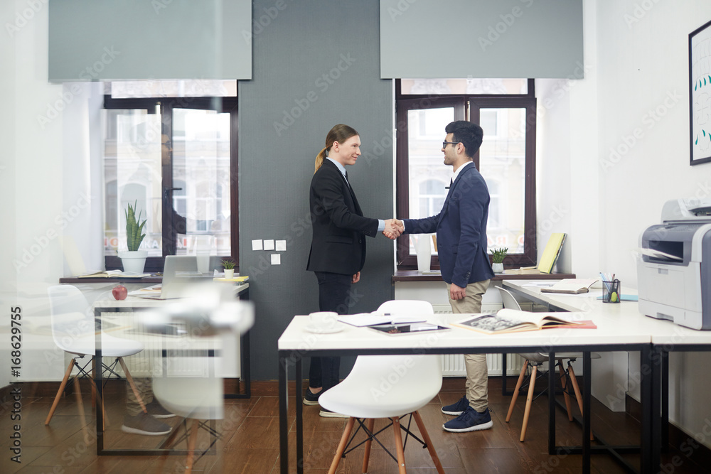 Profile view of young business partners shaking hands while standing at modern open plan office, they smiling at each other as sign of respect