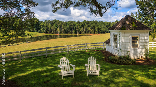 Valokuva 2 Adirondack chairs sit on a lawn in front of a white fence under a broad tree