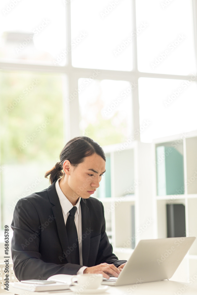 Confident Asian businessman wearing classical suit sitting in front of laptop and working on promising project, interior of modern office with panoramic windows on background Confident Asian businessman wearing classical suit sitting in front of laptop and working on promising project, interior of modern office with panoramic windows on background