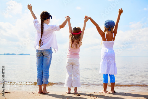 Back view of three ecstatic girls holding by hands while raising them in front of the sea