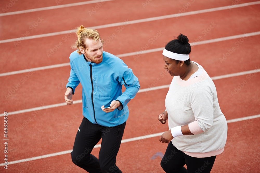 Obraz premium Young instructor and plump female jogging together on stadium racetrack before marathon competition