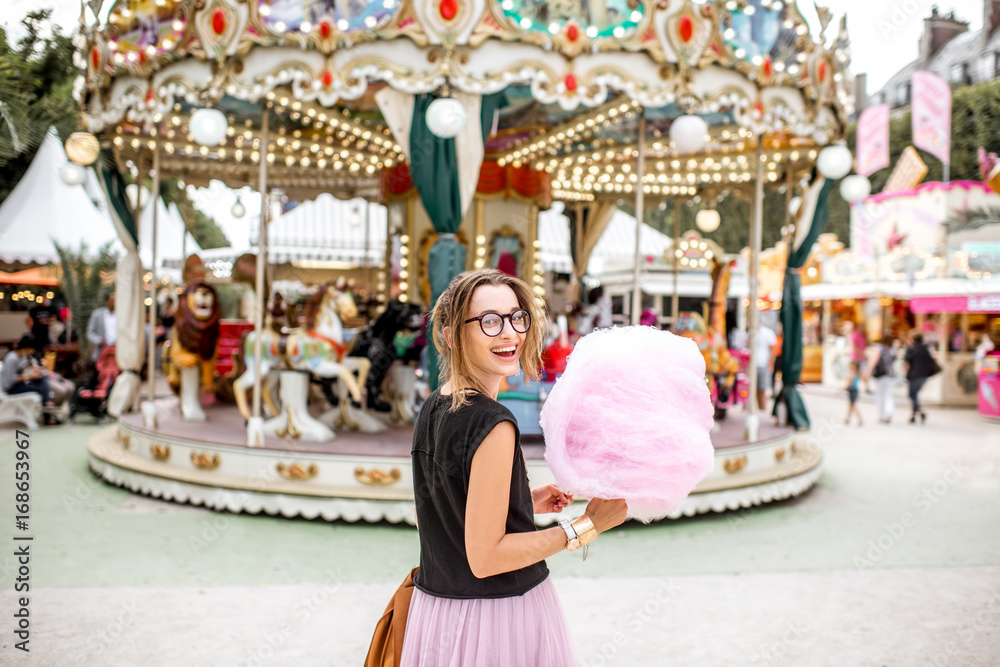 Young woman standing with pink cotton candy outdoors in front of the carrousel at the amusement park
