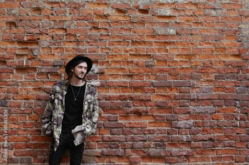 Horizontal lifestyle portrait of happy trendy looking young male hipster posing outdoors against red brick wall background with copy space for your content, wearing hat, jeans and camouflage bomber