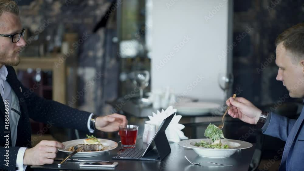 workers discussing project with tablet eating in cafe during business lunch. Blond man with beard in white shirt, black jacket, spectacles speaking, explaining issues, holding moving pictures on workers discussing project with tablet eating in cafe during business lunch. Blond man with beard in white shirt, black jacket, spectacles speaking, explaining issues, holding moving pictures on