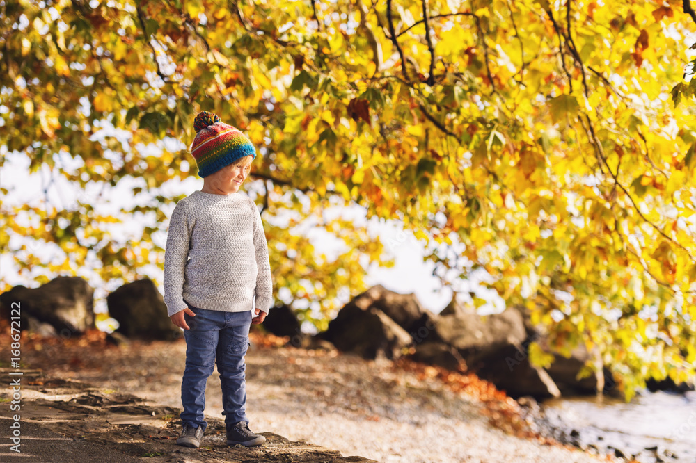 Fototapeta premium Little boy playing by the lake on a sunny day, wearing warm hat, grey knitted pullover, trousers and shoes