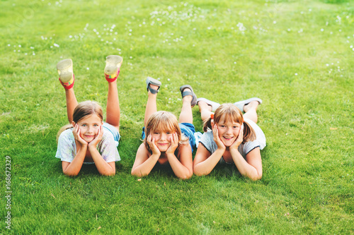 Group of three happy kids lying on fresh green grass, looking at camera