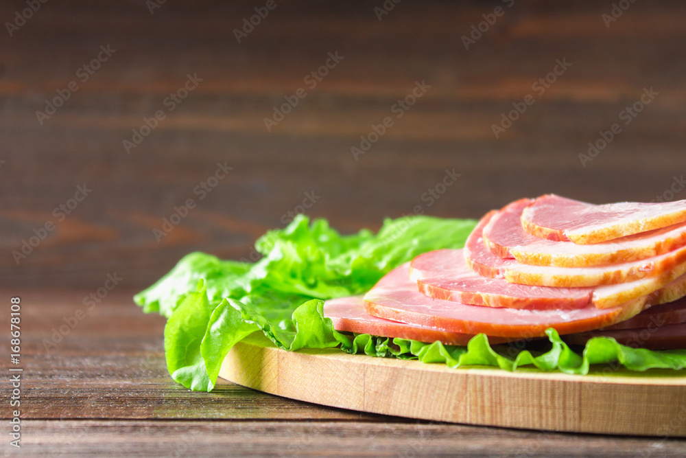 Sliced ham with fresh green lettuce leaves on a round cutting board. Meat products on a brown wooden table.