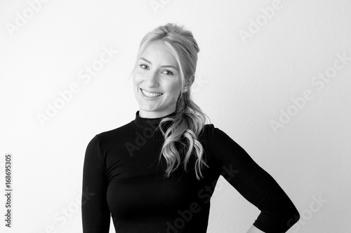 Zufriedene junge Frau mit blonden langen wehenden Haaren im schwarzen engen Pullover vor einer weißen Wand in reduzierter Optik 