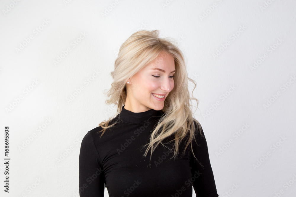 Zufriedene junge Frau mit blonden langen wehenden Haaren im schwarzen engen Pullover vor einer weißen Wand in reduzierter Optik Zufriedene junge Frau mit blonden langen wehenden Haaren im schwarzen engen Pullover vor einer weißen Wand in reduzierter Optik