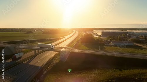Aerial View of White Semi Truck with Cargo Trailer Passing Highway Overpass/ Bridge. Eighteen Wheeler is New, Loading Warehouses are Seen in the Background.