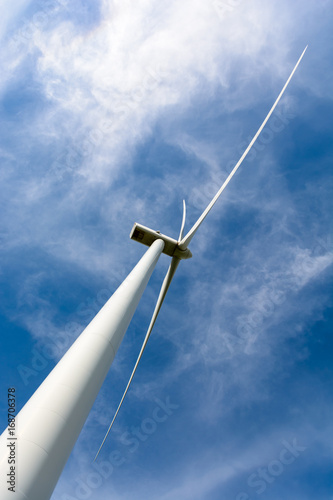 Low angle and diagonal view of a wind turbine against blue sky with fluffy white clouds.