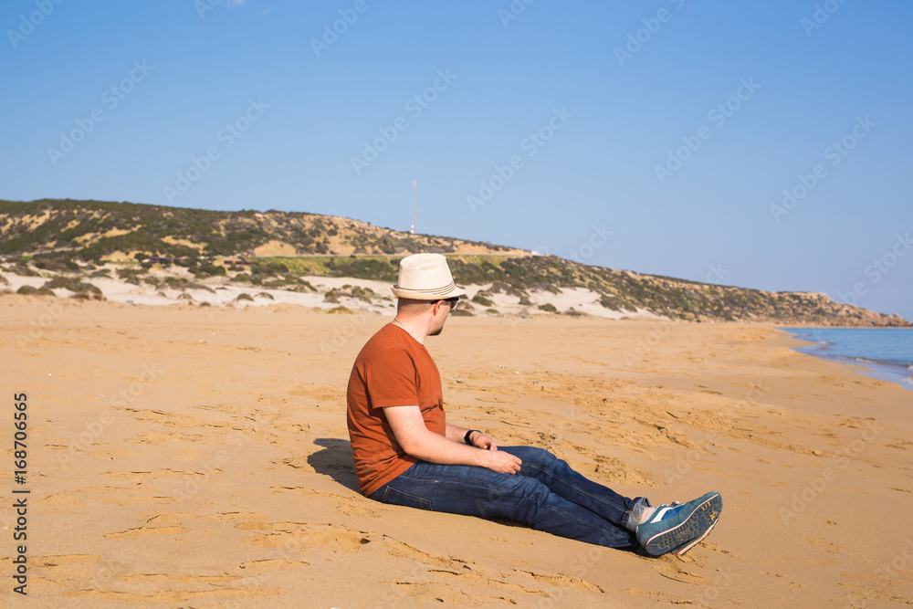 a man sitting on the beach in pants and a brown t-shirt, watching the sea
