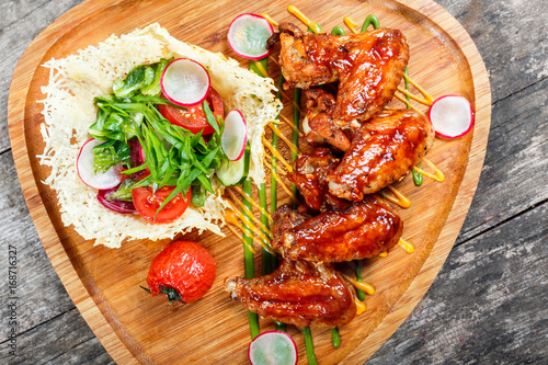 Fried chicken wings with fresh salad, grilled vegetables and bbq sauce on cutting board on wooden background close up. Hot Meat Dishes. Top view