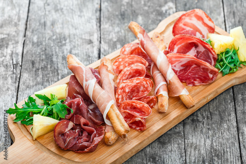Antipasto platter cold meat plate with grissini bread sticks, prosciutto, slices ham, beef jerky, salami and arugula on cutting board on wooden background. Meat appetizer. Top view