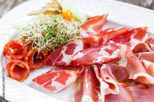 Antipasto platter cold meat plate with prosciutto, slices ham, salami, decorated with physalis and slices of melon on wooden background. Meat appetizer. Top view