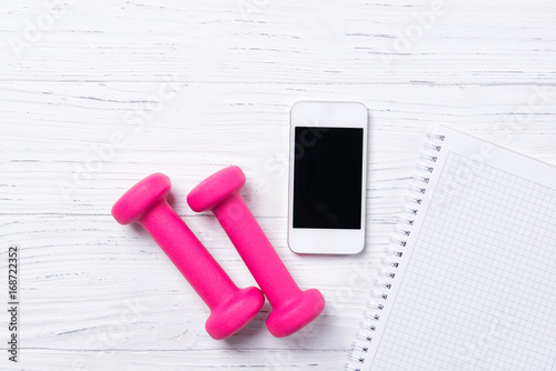 Fitness concept, pink dumbbells, smart phone and notebook on wooden background, top view with copy space