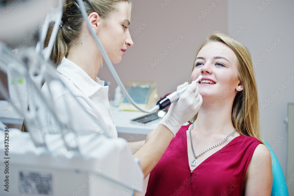 Attractive patient in red-violet dress laying on the dental chair while female dentist treating her teeth with special instruments. Attractive patient in red-violet dress laying on the dental chair while female dentist treating her teeth with special instruments.