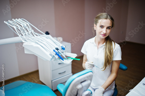 Good-looking female dentist posing with some dental instruments in her hand in white coat in a modern well-equipped cabinet.