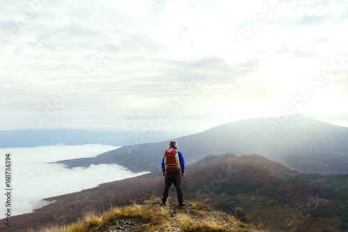Back view of young tourist hiker with backpack standing on the top of the mountain and looking at beautiful yellow autumn landscape sunset over clouds