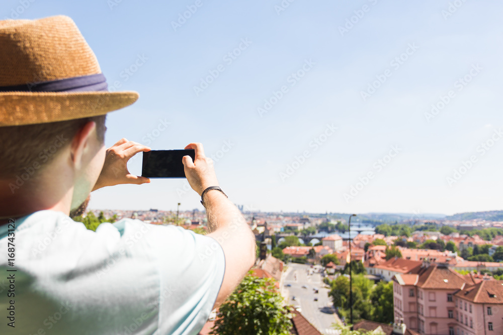 Fototapeta premium Back view of a young man tourist taking photo with smartphone in front of city