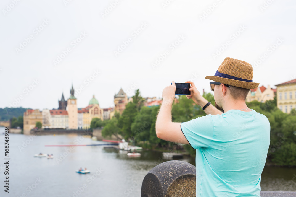 Back view of a young man tourist taking photo with smartphone in front of river and city