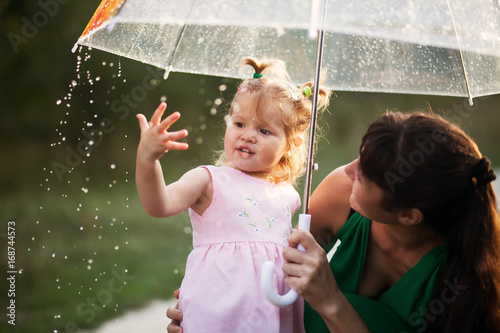 Happy funny family with umbrella under the autumn shower. Girl and her mother are enjoying rainfall. Kid and mom are playing on the nature outdoors.