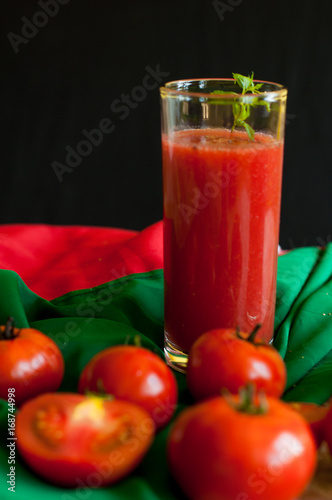 Glass and bottle of tomato juice with vegetables and green napkin on wooden background