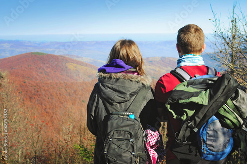 Young woman and man standing with their backs turned, looking at mountain range covered in red, orange, yellow deciduous forest under blue cloudless sky on warm fall day. October, Carpathians, Ukraine