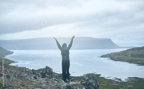 Girl in warm clothing stands on the cliff on background of mountains and fjords of Iceland. woman stands with her hands up