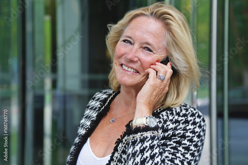femme senior active et souriante téléphonant devant centre d'affaires avec buildings 