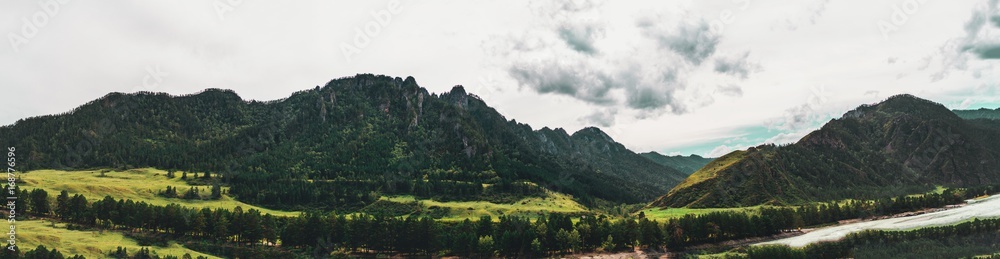 Panoramic image of Altai mountain-ridge near Katun river on summer overcast day: grass meadows, forests on flanks of hills, river coastline, peaks and white cloudy sky, Russia