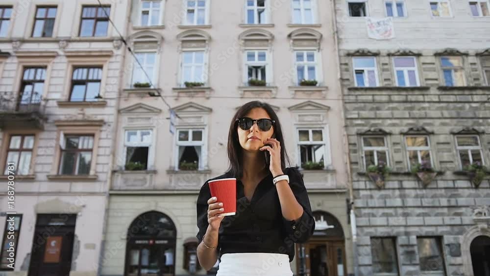 A young business woman in the glasses goes around the city,talks on the phone and drinks coffee A young business woman in the glasses goes around the city,talks on the phone and drinks coffee