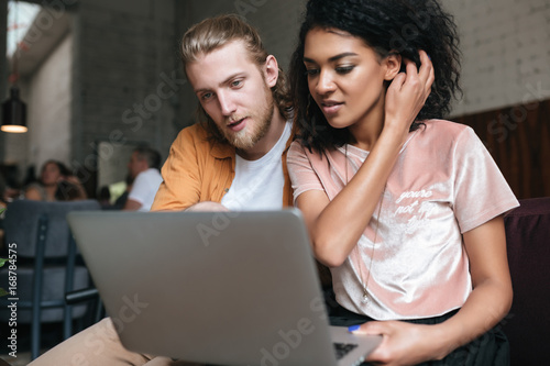 Portrait of young man and girl sitting in restaurant and working on laptop. Nice African American girl with dark curly hair sitting with friend at cafe while looking at laptop and tidying her hair