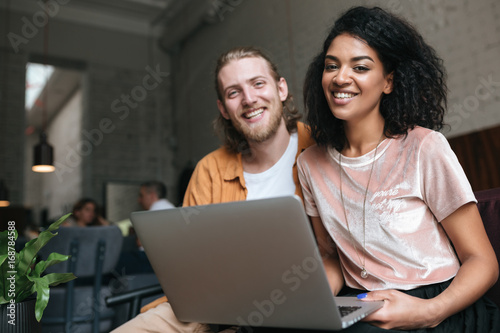 Portrait of young man and girl sitting in restaurant and happily looking in camera with laptop in hands. Nice African American girl sitting with friend at cafe. Boy and girl happily looking at laptop