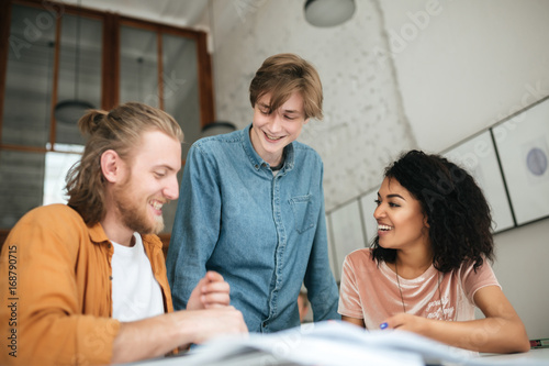 Portrait of young cheerful people discussing something in office. Two boys with blond hair and girl with dark curly hair working together on new project in classroom. Cool students studying together